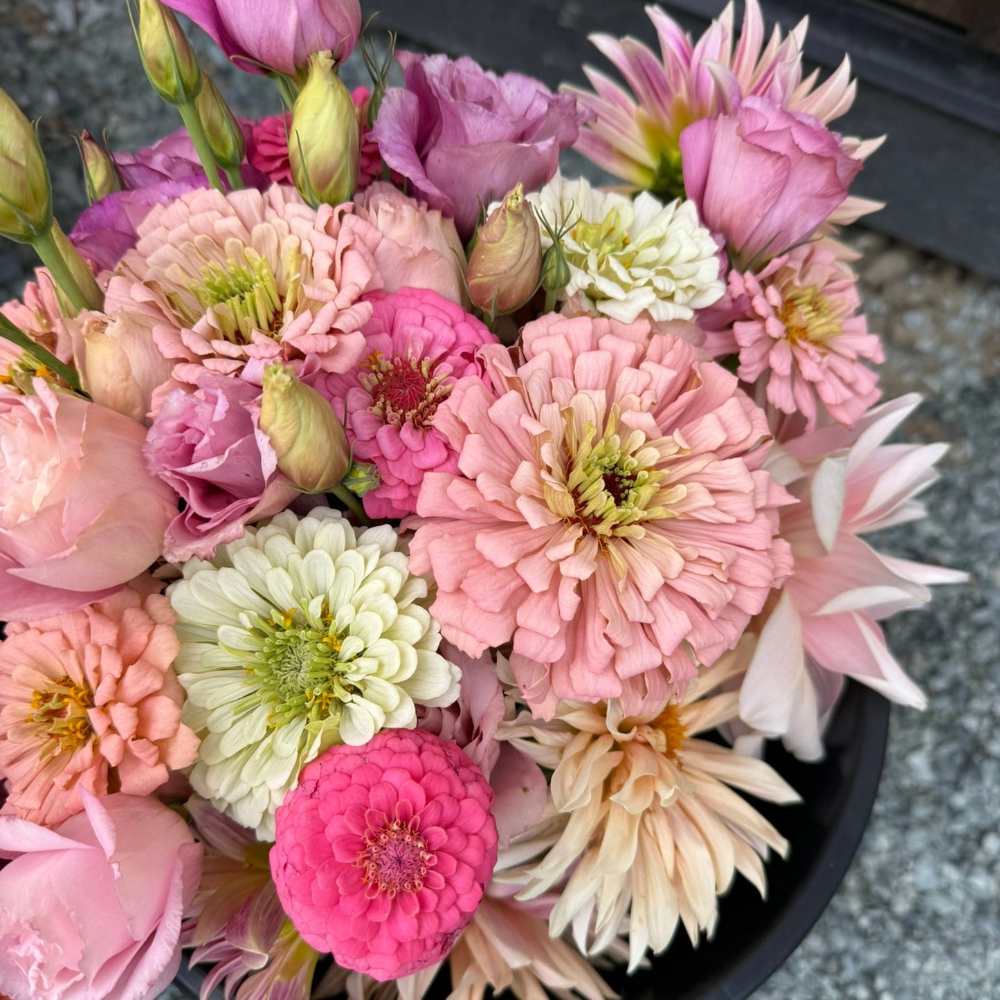 Bouquet of pink, white, and green flowers in a black pot on a gray pavement.